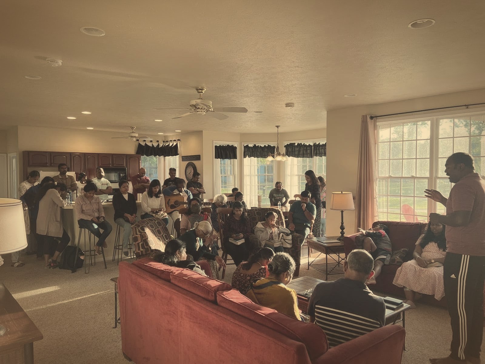 Church members gathered in a sunlit living room with Bibles, praying together as a leader speaks