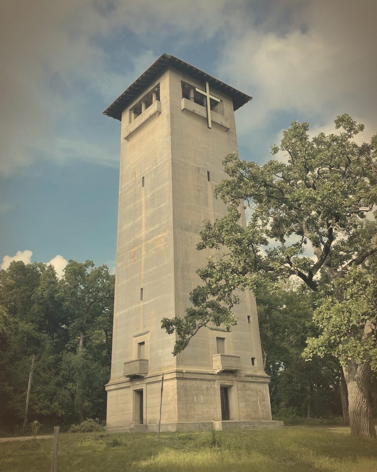 A tall stone bell tower with a cross near its top, rising above green trees against a blue sky