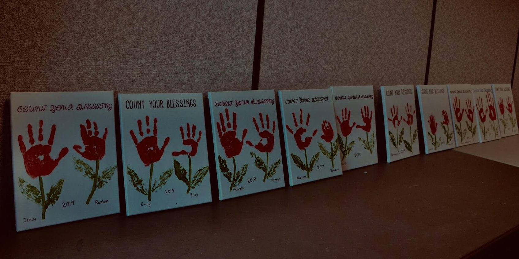 A row of canvases painted by the children, each with red handprint flowers, their names and the words Count Your Blessings