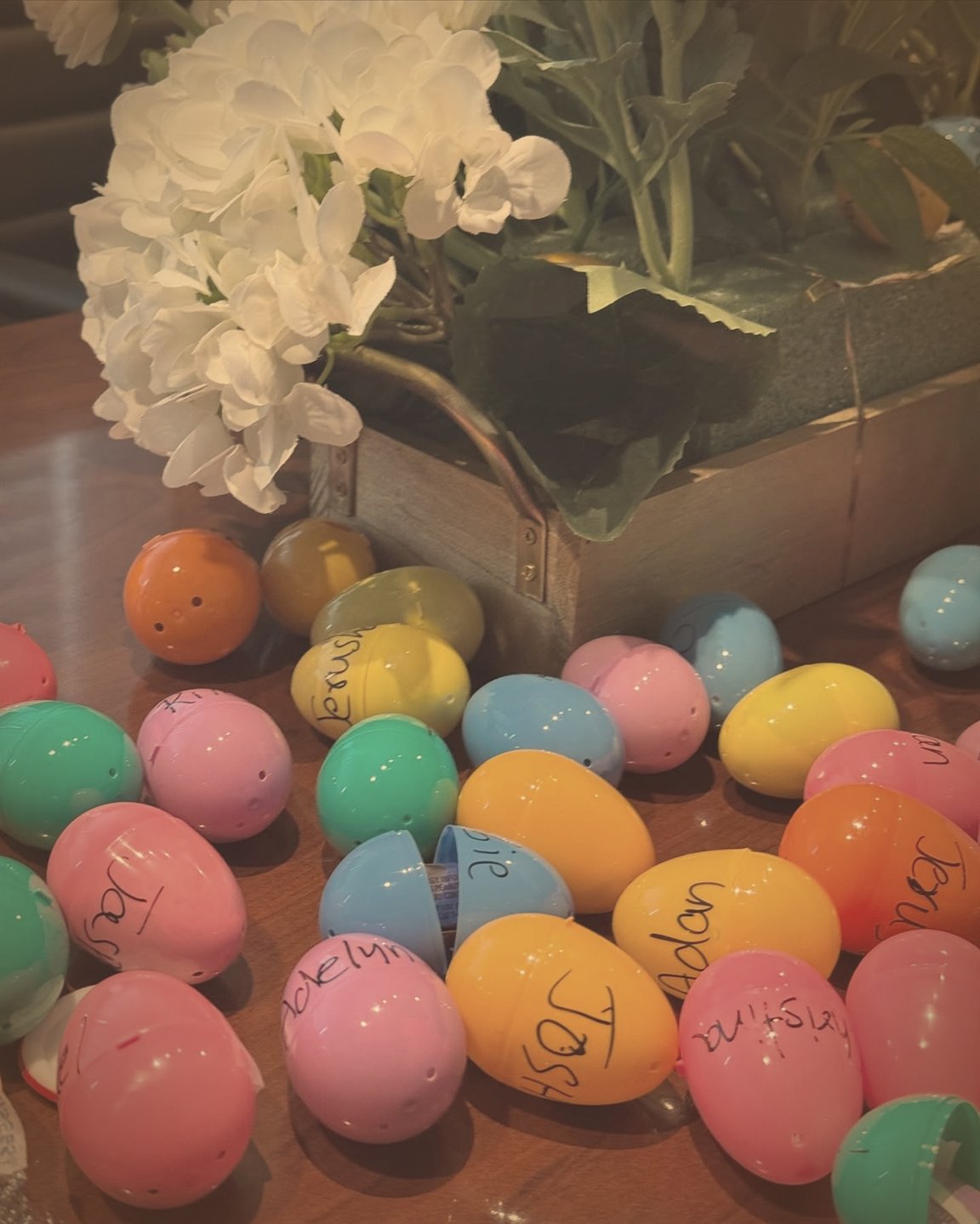 Colourful Easter eggs, each with a child's name handwritten on it, spread on a table beside white flowers