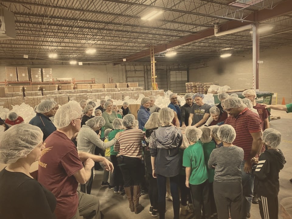 Church volunteers in hairnets gathered around a packing table at a Feed My Starving Children warehouse, praying together before packing meals