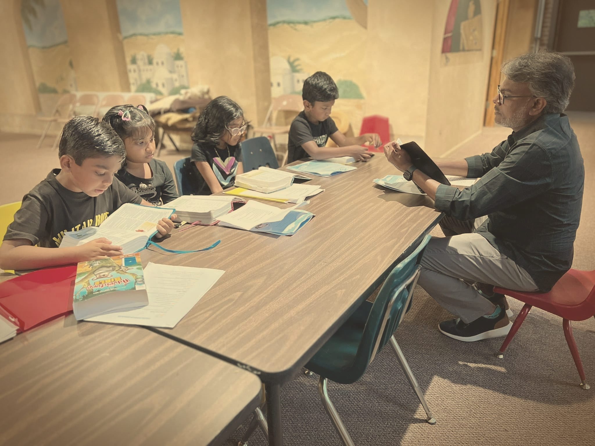 A Sunday School teacher reading Scripture with children as they follow along in their Bibles at a classroom table