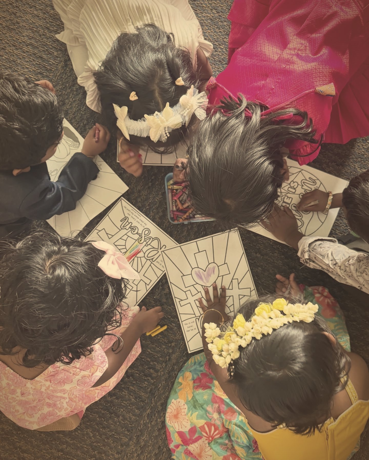 Children in festive clothes and flower hair garlands gathered on the floor colouring 'He is risen' Easter pages